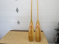 Photo showing both wooden paddles standing against a white garage door on a wooden surface, full length visible.