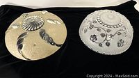 Two glass ceiling light shades displayed side by side on black fabric. One is amber with silver swirl accents; the other is clear with floral raised reliefs.