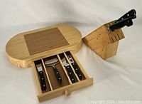 Photo showing the wooden cheese board with a darker square center, and the inset drawer pulled out revealing four cheese cutting utensils with black handles.