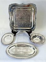 Four silver-plate serving trays consisting of square filigreed, oval etched, round serving dish, and small oval tray with slight pitting shown together on white background.