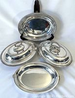 Four vintage silver-plate serving trays displayed on stand. Two trays with lids and handles, two simple platters. All in reflective silver-plate finish with beaded edge decoration.