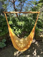 Full view of yellow woven hammock chair hanging outdoors with wooden bar and green foliage background.