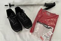 Photo of all items: black Hoka Clifton 6 running shoes, packaged red Saucony sleeveless shirt, and silver Komerdell hiking pole.