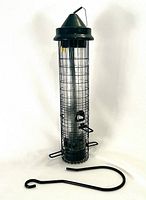 Full view of black metal cage bird feeder standing upright with hanging hook laying flat beside it on white background.