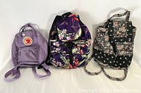 Three backpacks in a row, left to right: mauve Fjallraven Kanken, purple floral Nani Island, and black/pink olive floral soft-sided backpack.