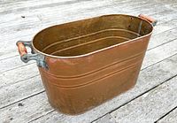 Vintage copper wood bin with wooden handles on the sides, showing oval shape and copper patina.