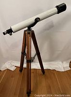 Full view of vintage white telescope mounted on wooden tripod with extending legs against plain background.