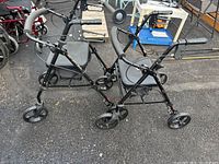 Two black 4-wheel rollators side by side on pavement, showing seats and handles with brakes.