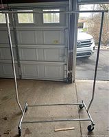 Full view of collapsible metal clothing rack on wheels in garage with some rust on base.