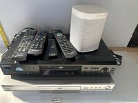 Photo showing Sony DVD player stacked on Panasonic DVD recorder with multiple remote controls and a white Sonos speaker on top.