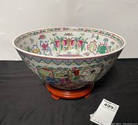 Front and side view of large Asian decorative porcelain bowl with colorful hand-painted traditional scenes and floral patterns on wooden stand.
