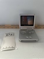 Game Boy Advance SP handheld gaming console open, showing screen and buttons with a ruler and lot tag next to it.