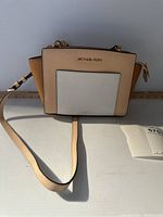 Front and side view showing the beige, tan, and white color blocking and gold hardware of the Michael Kors handbag, including the front slip pocket and attached shoulder strap.