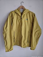 Front view of medium yellow MEC rain jacket hanging against white wall, showing full zipper, hood and two zippered pockets.
