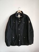 Front view of black men's Fjallraven jacket with four flap pockets and snap buttons hanging on hanger.