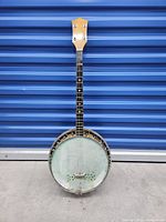 Front view of vintage 1920s Harmony banjo showing painted skin head and wooden neck against a blue background.
