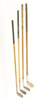 Four antique golf clubs standing upright showing wooden shafts, various grip types, and metal club heads of different styles and lengths.