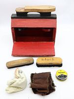 Photo of open red wooden shoe shine box showing the drop front opened revealing inside compartments and included items: two brushes, shoe polish tin, and cloths.