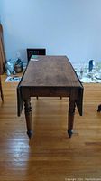 Front view of antique pine drop leaf table with leaves down, showing turned legs and wooden surface with visible scratches.