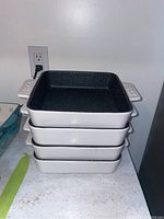 Stack of four square baking dishes with black speckled enamel interiors and white exteriors, stacked on a countertop.