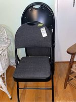 Set of three black metal folding chairs with dark gray upholstered seats and backs, stacked together showing full chairs front and side views.