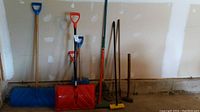 Photo showing all garden tools including three winter shovels, metal rake, sledgehammer, leaf pactor and broom arranged against a wall.