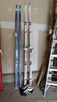 Full view of two pairs of cross country skis standing upright, blue and white skis with bindings are visible along with black and white ski boots placed in front of the skis.