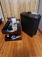 Overall view showing black Shredmaster paper shredder and two wooden organizer drawer trays filled with supplies placed on wood floor against wall.