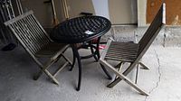 Two weathered teak folding chairs and one round lattice metal outdoor table arranged together showing weathered condition and outdoor use wear.