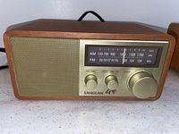 Front view of the Sangean AM/FM radio showing wooden casing, speaker grille, analog dial with AM/FM frequencies, and control knobs.