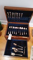Overview of flatware set in oak case with velvet interior. Visible forks, spoons, and other utensils arranged in holders inside case and tray.
