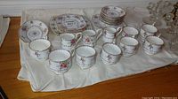 Photo showing 8 teacups and saucers, 4 side plates, luncheon plate, serving plate, a dish, 3 creamers and a sugar bowl arranged on a cloth.