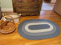 Three items together: oval braided rug in blue, gray, cream shades, small round wicker basket with handle, larger oval wicker basket with white rope handles.