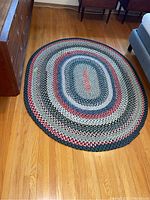 Photo showing the entire oval braided rug placed on a hardwood floor next to furniture including a dresser and a chair.