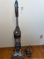 Front view of Hoover cordless vacuum cleaner standing upright on wooden floor against wall, showing handle, dust container and base.