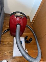 Red Miele S2 Contour vacuum cleaner with hose, wand, floor attachment, and replacement bags on hardwood floor near wall.