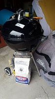 Full image of Weber black kettle charcoal grill on tripod base with wheels, unopened bag of Kingsford charcoal placed in front, grey Napoleon barbeque cover partially visible behind