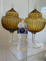 Pair of vintage Italian Empoli amber glass pedestal jars with matching lids. Each jar has a ribbed bowl sitting on a clear twisted stem pedestal and is topped with a pointed finial lid. A water bottle is placed beside for scale showing jars are about twice the height of a standard water bottle.