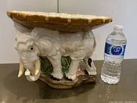 Side view of the vintage glazed bisque elephant pedestal next to a water bottle for scale.
