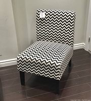 Accent chair upholstered in black and white zig zag pattern fabric, four dark wooden legs, standing on dark wooden floor against light-colored wall