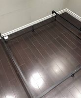 Photo shows black metal adjustable bed frame assembled on dark wood floor. Frame is rectangular with three support legs visible including center leg.