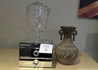 Photo showing a large clear glass apothecary jar, a ceramic vase with geometric patterns, and a mirrored jewelry box with crystal knobs stacked together on a surface.