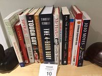 Photo showing a collection of 11 assorted books with visible titles and authors, placed vertically on a surface between two objects.
