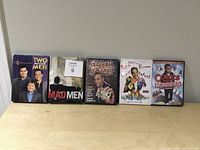 Photo showing 5 DVD cases together on a light wood surface against a plain wall: Two and a Half Men Season 4, Mad Men Season 1, and three Russell Peters DVDs including 'Red, White and Brown' and 'Comics Without Borders'.