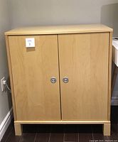 Front view of light birch veneer cabinet with two doors and circular metallic handles, standing on block-style legs.