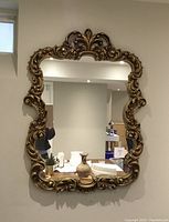 Photo of a large rectangular mirror with a heavily ornate gold frame featuring intricate carvings and scrollwork, hanging on a wall reflecting some home interior.