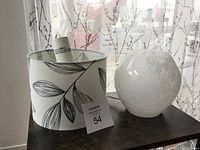 Photo showing a hanging pendant light shade with botanical leaf pattern next to a small white floral glass table lamp on a dark surface with a window and sheer curtains in the background.
