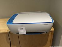 White and blue HP DeskJet 3632 printer placed on a wooden cabinet, showing the top and front views with power cord attached.