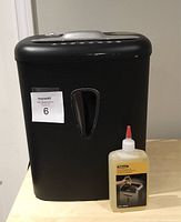 Full front view of the black Amazon Basics shredder with control panel on top and bin window, next to a bottle of shredder oil lubricant.