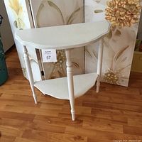 Small vintage side table painted white with semi-circular shape and turned legs, two-tier with a lower corner shelf, placed on a wood floor with decorative paintings behind.